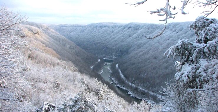 Lightly snow-covered landscape of a gorge with a winding river and steep hillsides. Snow on tree branches in foreground. Mostly cloudy sky.