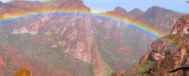 Desert mountain valley with a rainbow arcing below a mostly clear blue sky. Light rain falls on the dry brown ground where scattered desert plants grow.