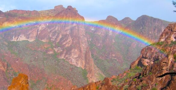 Desert mountain valley with a rainbow arcing below a mostly clear blue sky. Light rain falls on the dry brown ground where scattered desert plants grow.