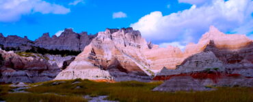 Dramatic landscape with sharply eroded cliffs and pointed rock formations, some in shadow. The foreground is covered in tall grass. A clear blue sky with patches of white, fluffy clouds completes the scene.