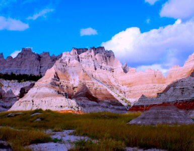 Dramatic landscape with sharply eroded cliffs and pointed rock formations, some in shadow. The foreground is covered in tall grass. A clear blue sky with patches of white, fluffy clouds completes the scene.