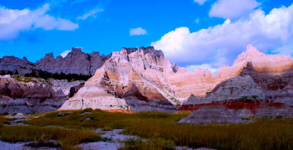 Dramatic landscape with sharply eroded cliffs and pointed rock formations, some in shadow. The foreground is covered in tall grass. A clear blue sky with patches of white, fluffy clouds completes the scene.