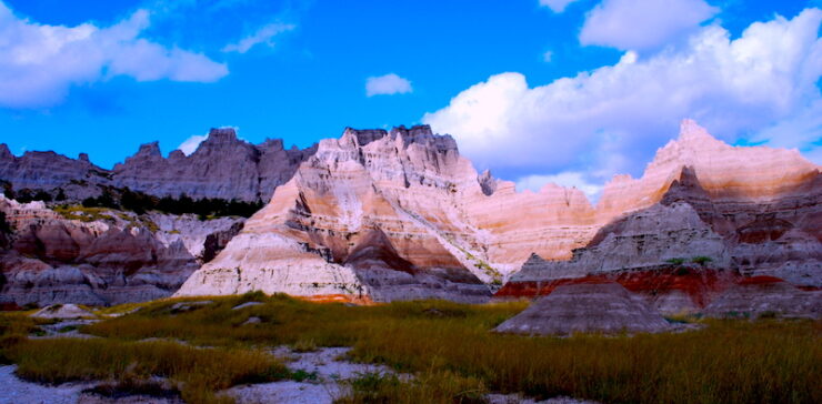 Dramatic landscape with sharply eroded cliffs and pointed rock formations, some in shadow. The foreground is covered in tall grass. A clear blue sky with patches of white, fluffy clouds completes the scene.