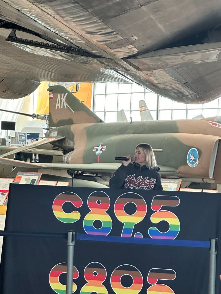 An image of Anabel Englund behind a DJ booth singing into a mic with the c895 logo and a jet behind her