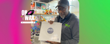 A person in a cap and sweater holds a vinyl record album labeled "Test Pressing" in a vibrant, colorful store filled with vinyl records and music paraphernalia, with a neon sign saying "ON WAX" visible in the background.