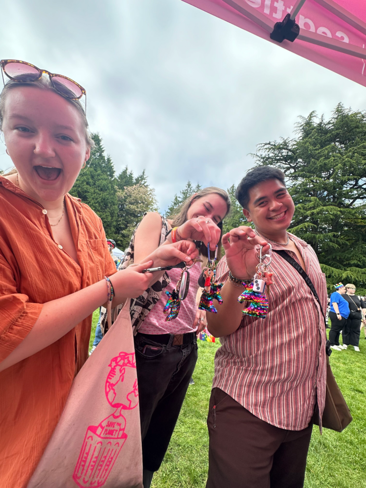 Three individuals at an outdoor event, smiling and holding up colorful beaded keychains towards the camera. The keychains feature vibrant, multi-colored beads and charms. One individual wears orange attire and sunglasses on their head, another is partially visible behind a pink umbrella, and the third sports a light shirt. A crowd and green trees are visible in the background, suggesting a lively, social atmosphere.