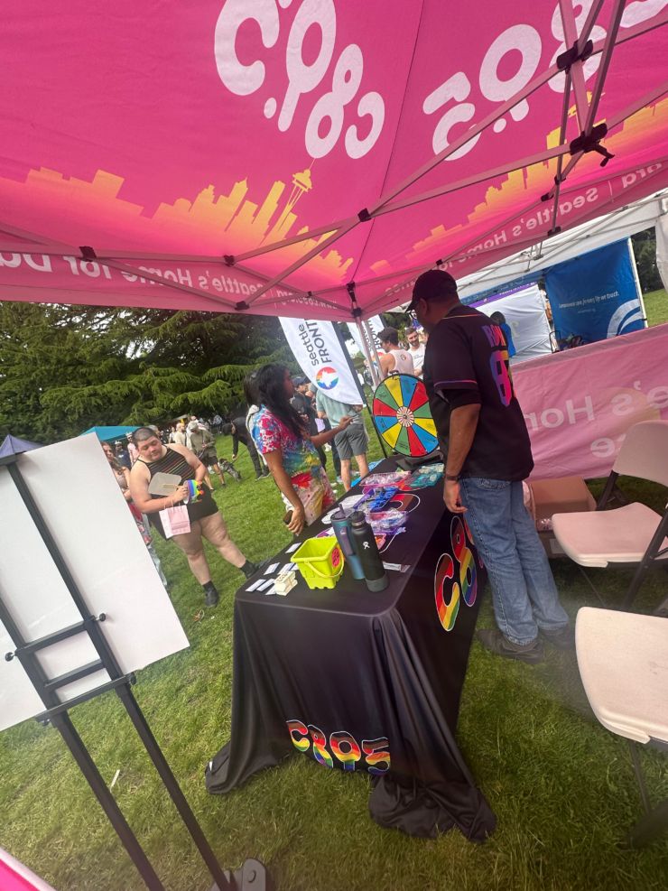 An outdoor event scene under a vibrant pink tent with "c895" printed on its canopy. An individual, wearing a dark backpack, is browsing a promotional table covered with a black cloth. The table displays an array of colorful disk-shaped items and other materials. To the left, another small white stand appears to hold additional information. People are visible in the background, engaging in event activities.