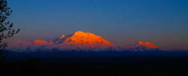A distant snow-capped peak under the diffused reddish sunlight, with soft atmospheric shadows, contrasting against a dark nighttime foreground.