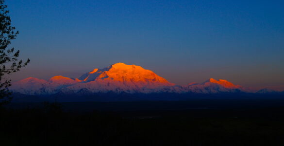 A distant snow-capped peak under the diffused reddish sunlight, with soft atmospheric shadows, contrasting against a dark nighttime foreground.