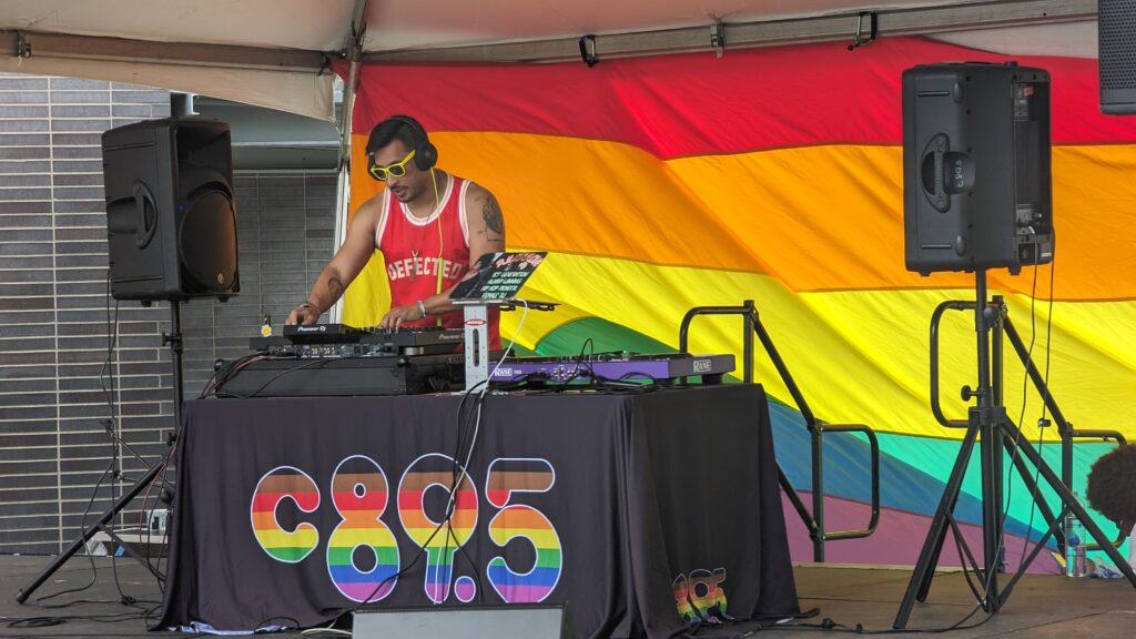 DJ Alfonso Tan standing in front of a Pride Flag while DJing on a stage with the c895 logo
