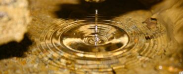 A clear pool of water inside a cave. A single drop disrupts the still surface, creating ripples. The bottom of the pool is covered in sediment and surrounded by brownish cave walls.
