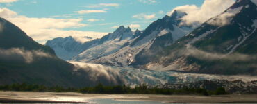 Glacier carving through valley to meet a body of water, surrounded by steep mountains with summer snow caps and evergreen trees along shore.