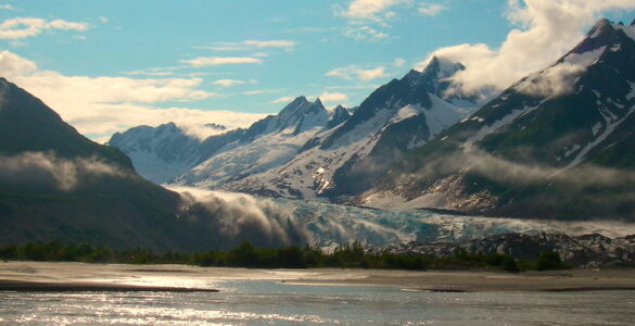 Glacier carving through valley to meet a body of water, surrounded by steep mountains with summer snow caps and evergreen trees along shore.