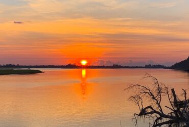 A sunrise casts an orange glow over a cloudy sky and a tranquil lake, with trees lining the shore.