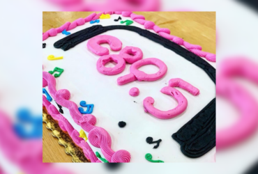 A birthday cake with pink and black frosting spelling out "c895", surrounded by colorful decorative elements.