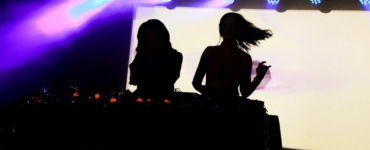 Silhouettes of two DJs performing at a music event with colorful stage lighting.