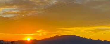 A sunset at the foot of a mountain, with its features darkened by shade. The clouds are glowing in bright orange.