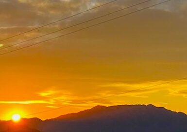 A sunset at the foot of a mountain, with its features darkened by shade. The clouds are glowing in bright orange.