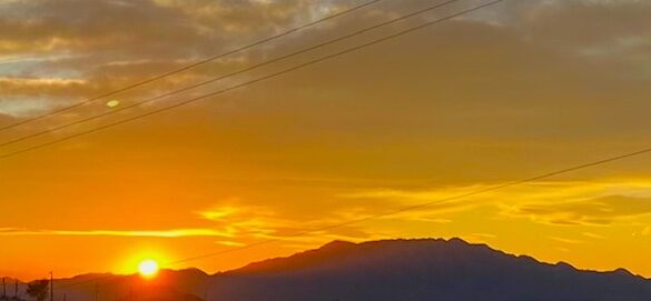 A sunset at the foot of a mountain, with its features darkened by shade. The clouds are glowing in bright orange.