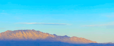 Sunrise light illuminates the mountain peak while the base remains in shadow, with dark blue tones. The sky above is blue with a few scattered clouds.
