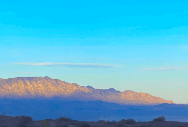 Sunrise light illuminates the mountain peak while the base remains in shadow, with dark blue tones. The sky above is blue with a few scattered clouds.