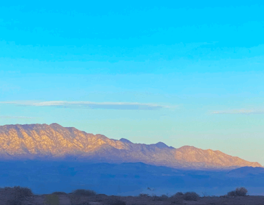 Sunrise light illuminates the mountain peak while the base remains in shadow, with dark blue tones. The sky above is blue with a few scattered clouds.