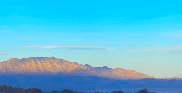 Sunrise light illuminates the mountain peak while the base remains in shadow, with dark blue tones. The sky above is blue with a few scattered clouds.