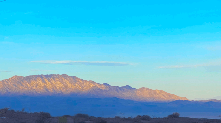Sunrise light illuminates the mountain peak while the base remains in shadow, with dark blue tones. The sky above is blue with a few scattered clouds.
