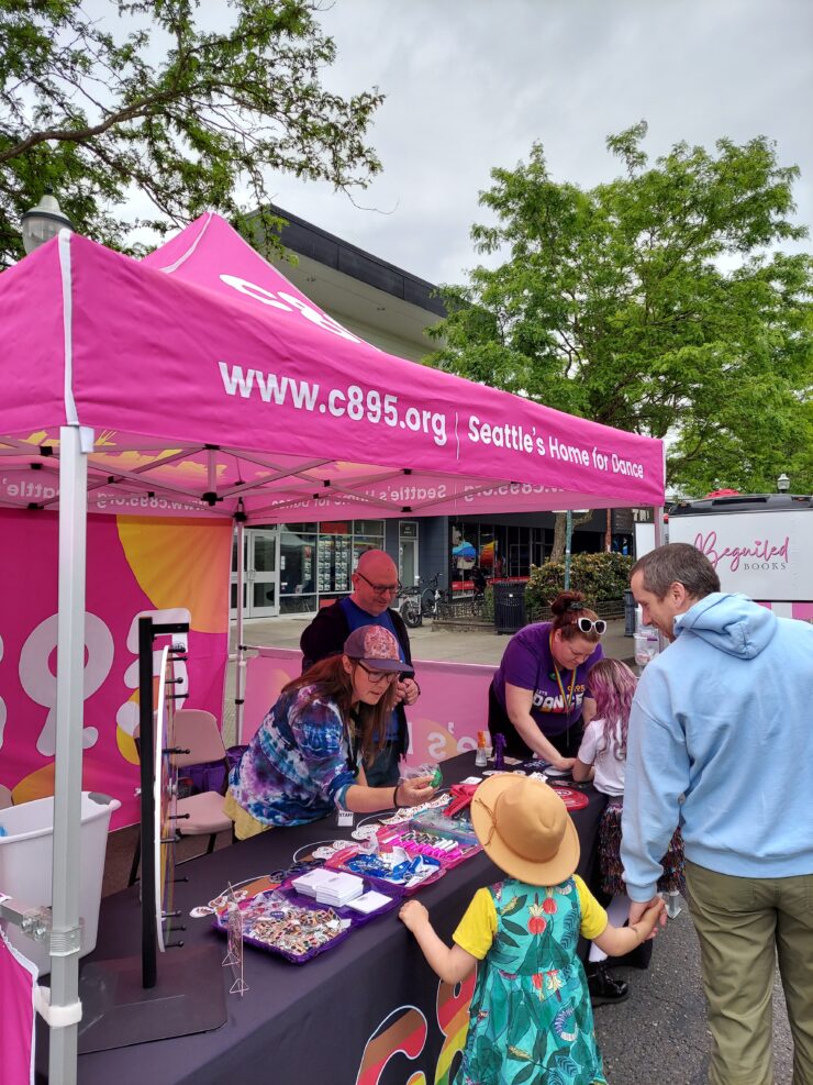 People visiting a promotional booth for C89.5, Seattle's Home for Dance, at an outdoor event. Attendees are interacting with staff and exploring merchandise on the table under a branded pink canopy.