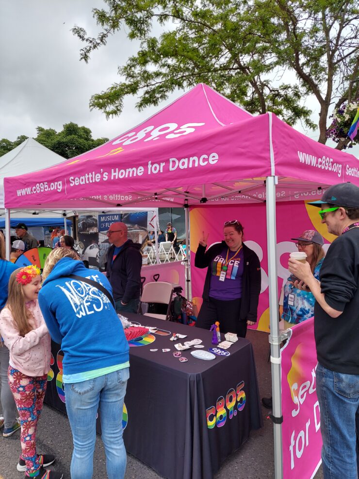 People visiting a promotional booth for C89.5, Seattle's Home for Dance, at an outdoor event. Visitors are interacting with staff and viewing promotional materials.