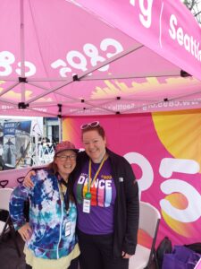 Two people smiling under a tent with a banner that reads "Let's Dance" and logos of C895 and Museum of Flight in the background. They are wearing casual clothing and event badges.