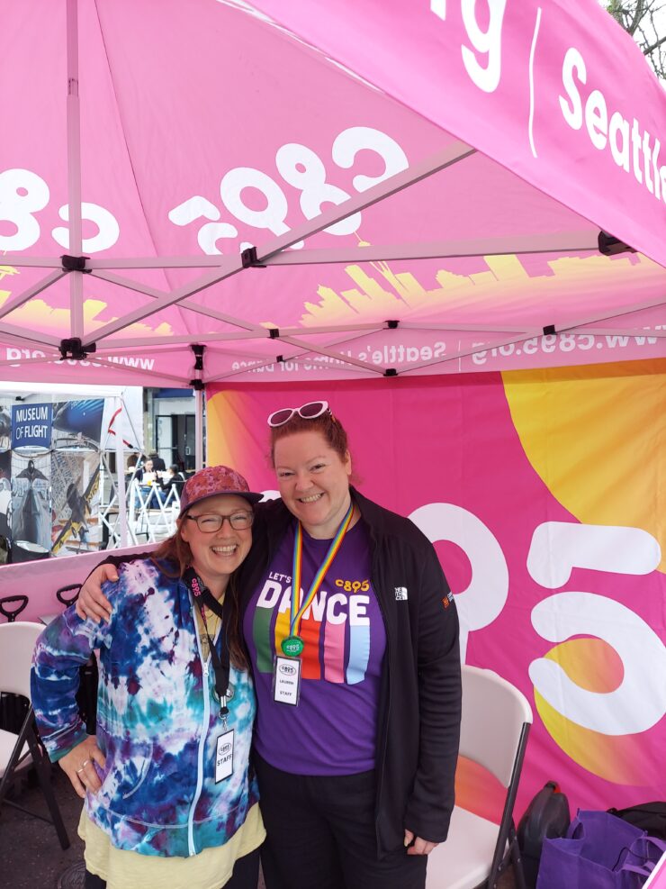 Two people smiling under a tent with a banner that reads "Let's Dance" and logos of C895 and Museum of Flight in the background. They are wearing casual clothing and event badges.