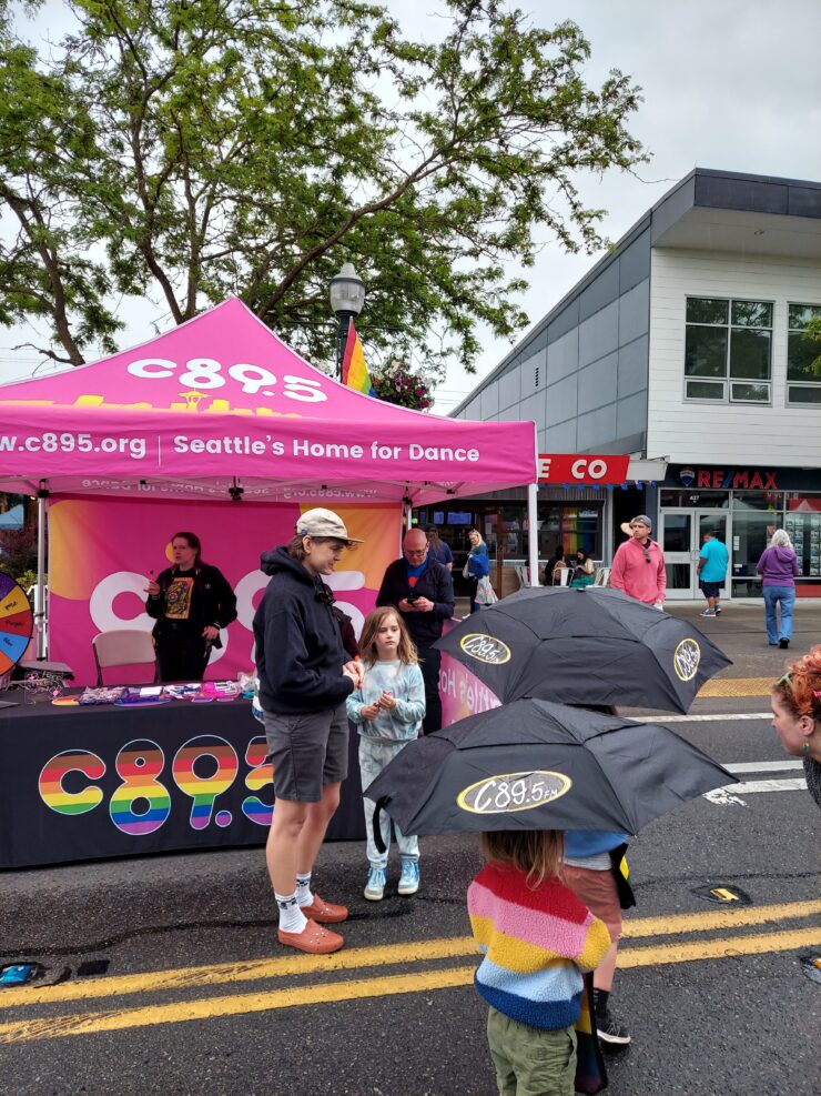 People gathered around the C89.5 booth at an outdoor event, with some holding umbrellas.