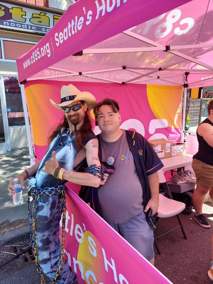 A person standing at a booth holding a small dog, both under a branded pink canopy. The person is wearing a blue top and multicolored pants, and the surroundings suggest a community event.