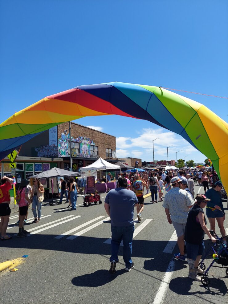 People gathered under a large rainbow arch at a street festival, with sunny weather and blue skies.