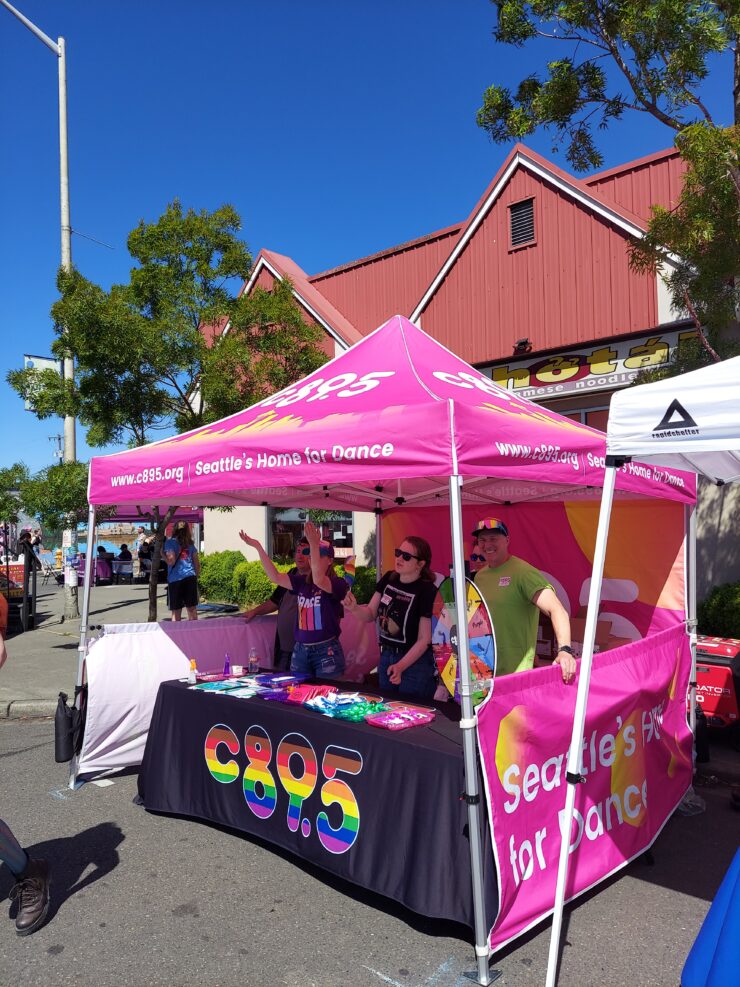 Individuals at a C89.5 radio station booth under a bright pink banner at an outdoor event. The booth features promotional materials and a colorful backdrop