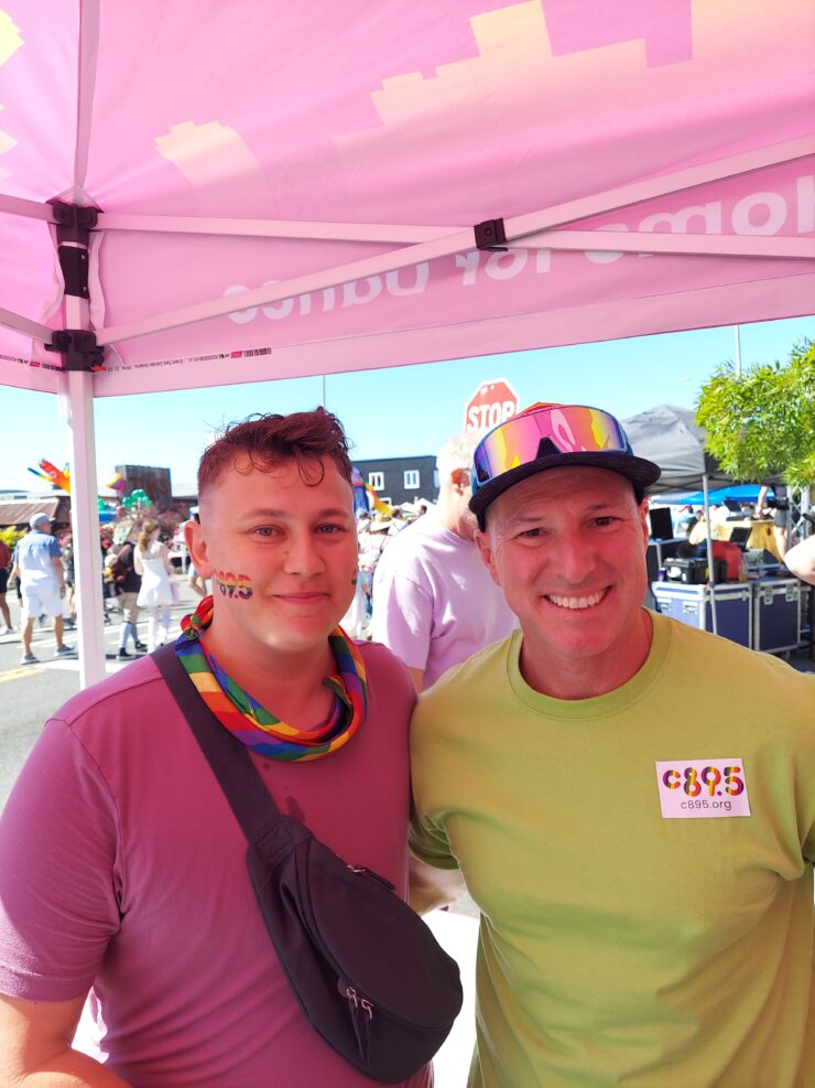 Two individuals standing under a pink canopy at an outdoor event, smiling at the camera, both wearing stickers and one with a rainbow ribbon.