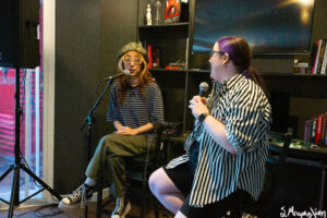 A person is performing music seated on a stool in a small room with recording equipment, while another person seated in the foreground appears to be singing into a microphone.