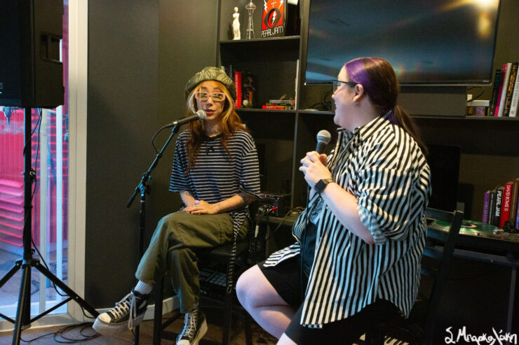 A person is performing music seated on a stool in a small room with recording equipment, while another person seated in the foreground appears to be singing into a microphone.