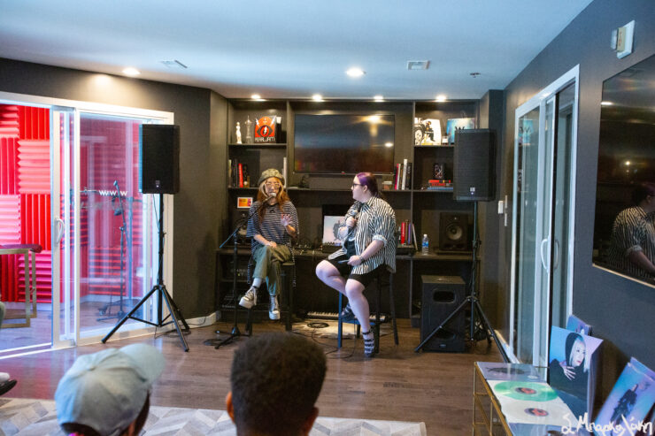 A person is performing music seated on a stool in a small room with recording equipment, while another person seated in the foreground appears to be singing into a microphone.