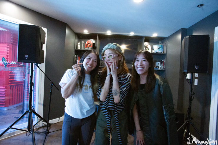 Three individuals smiling in a music studio, with one wearing a playful mask. Shelves with awards and a speaker are visible in the background.