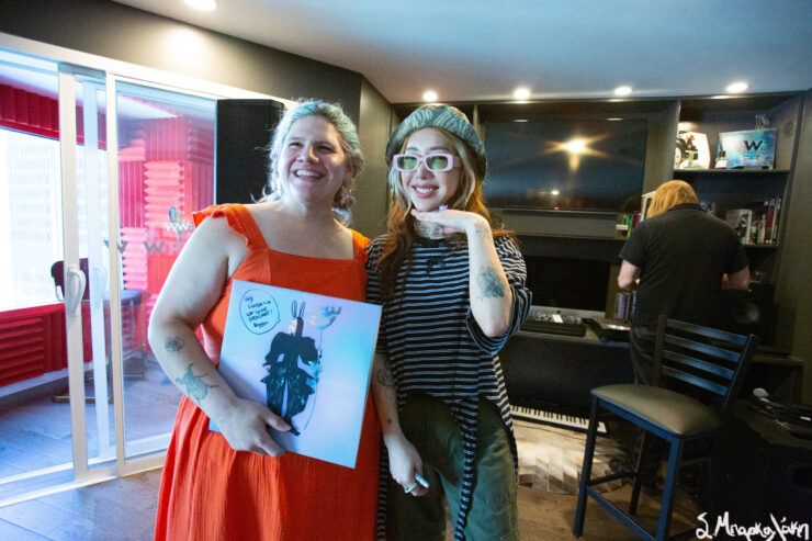 Three individuals smiling in a music studio, with one wearing a playful mask. Shelves with awards and a speaker are visible in the background.