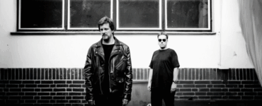 Two people standing against a brick wall under rectangular windows, one wearing a leather jacket and the other in a casual shirt and sunglasses.