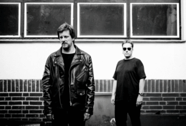 Two people standing against a brick wall under rectangular windows, one wearing a leather jacket and the other in a casual shirt and sunglasses.