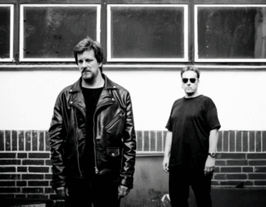 Two people standing against a brick wall under rectangular windows, one wearing a leather jacket and the other in a casual shirt and sunglasses.