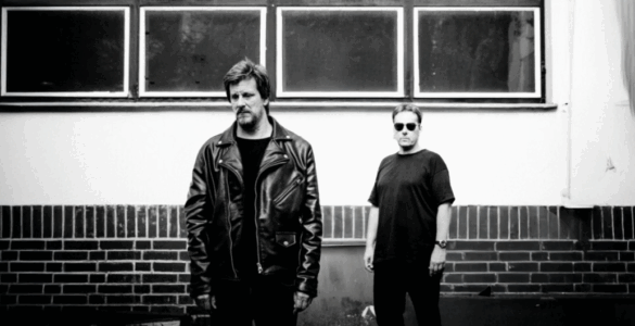 Two people standing against a brick wall under rectangular windows, one wearing a leather jacket and the other in a casual shirt and sunglasses.