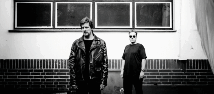 Two people standing against a brick wall under rectangular windows, one wearing a leather jacket and the other in a casual shirt and sunglasses.