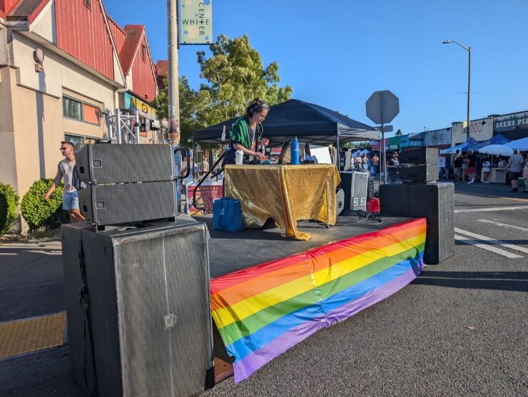 A musician performs on a small stage decorated with a rainbow pride flag in front of a casual outdoor audience at a street fair. There are tents and food trucks nearby.