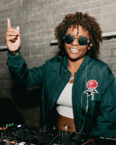 Person DJing at a club, raising one finger with a smile, wearing sunglasses and a jacket with a rose emblem.