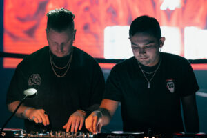Two DJs working at a mixing console in a club with vibrant lighting in the background.